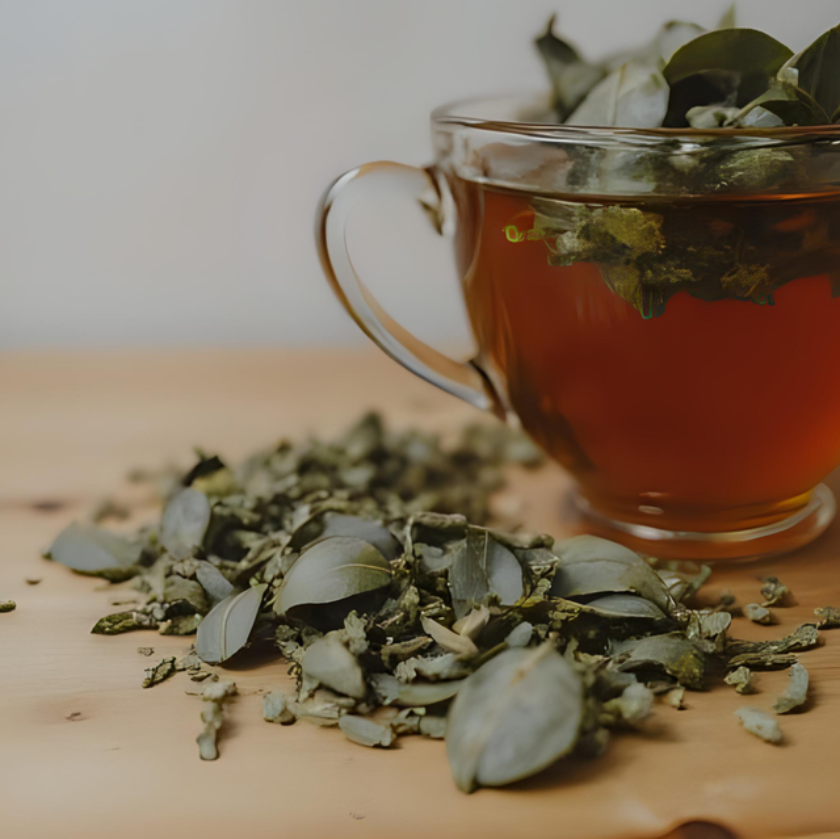A clear glass cup filled with amber tea and loose green leaves, set on a light wooden surface with scattered dried leaves in the foreground.